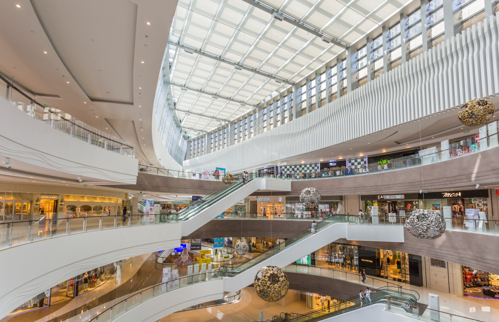 O interior de um shopping center moderno e espaçoso com vários andares, escadas rolantes, lojas, um teto de vidro e grandes luminárias esféricas penduradas.