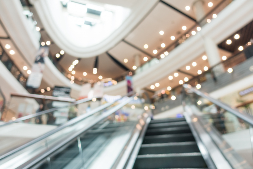 Interior desfocado de um shopping center moderno com escadas rolantes, múltiplos andares e luzes brilhantes no teto.