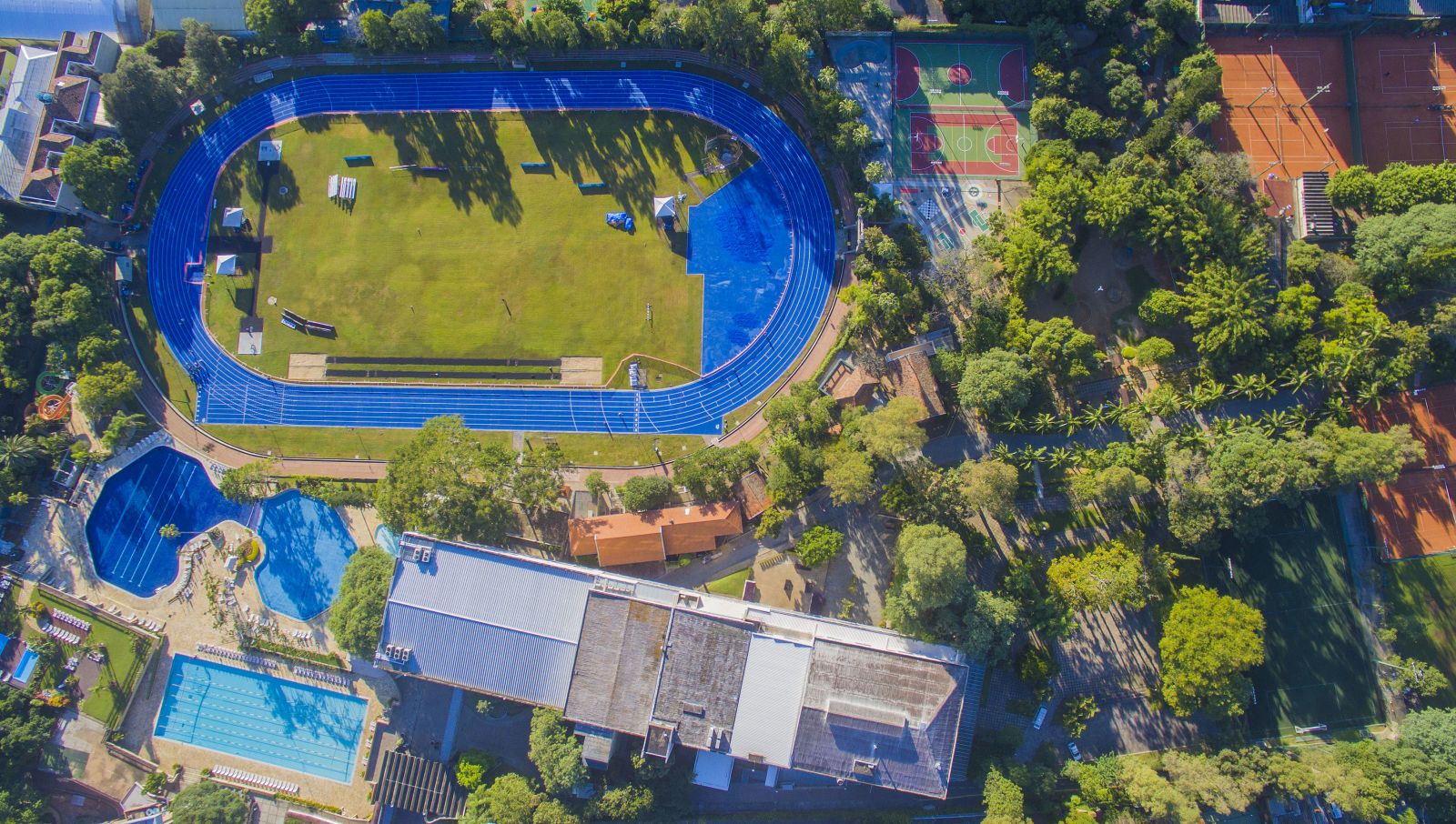 Vista aérea de um complexo esportivo com uma pista de corrida azul, campos verdes, várias piscinas, quadras de tênis e basquete, cercado por edifícios e árvores exuberantes.