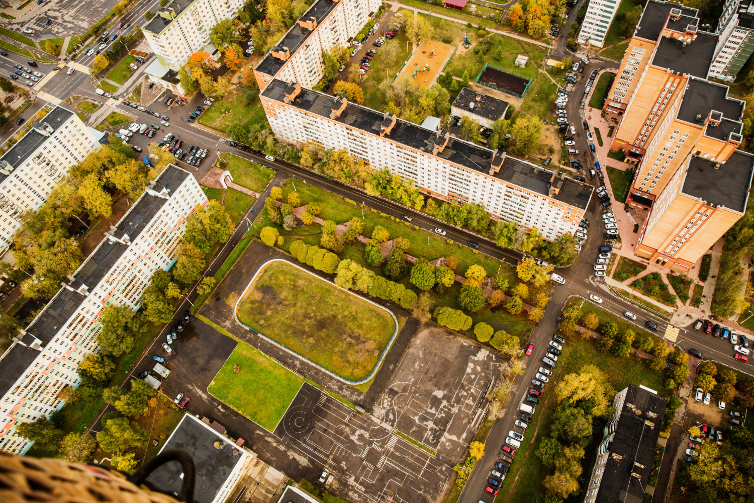 Vista aérea de um bairro residencial com grandes edifícios de apartamentos, muitas árvores com folhagem de outono, áreas verdes, ruas com carros estacionados e um campo esportivo.