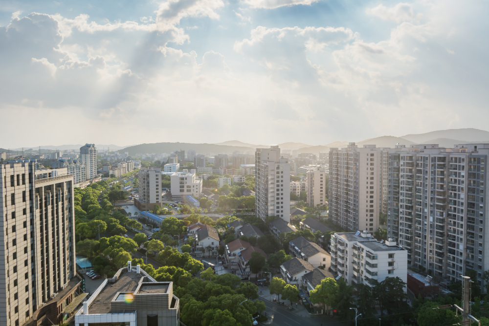 Vista aérea de uma cidade com muitos edifícios residenciais, ruas arborizadas e colinas ao fundo, sob um céu parcialmente nublado com raios de sol.