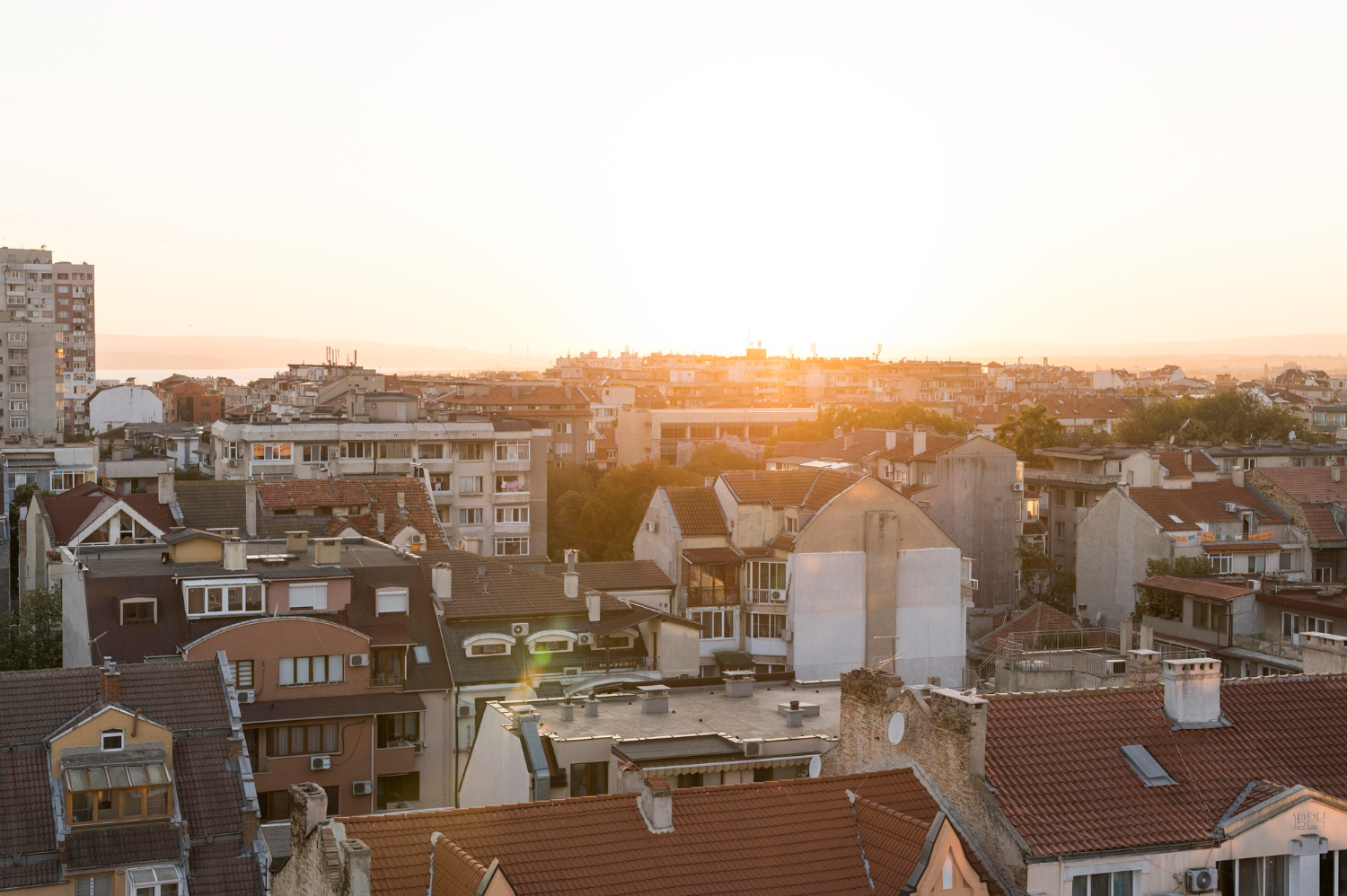 Vista aérea de uma cidade ao pôr do sol, com telhados de terracota e edifícios de vários andares, sob um céu laranja brilhante.
