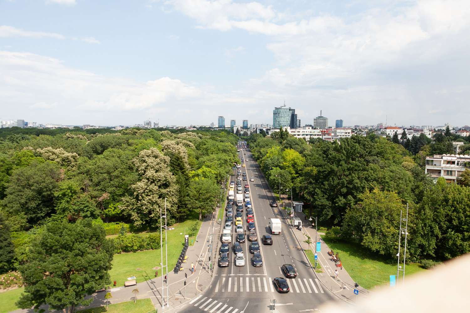 Vista panorâmica aérea de uma avenida movimentada com tráfego, ladeada por extensas áreas verdes e árvores, levando a uma paisagem urbana com edifícios ao fundo, sob um céu parcialmente nublado.