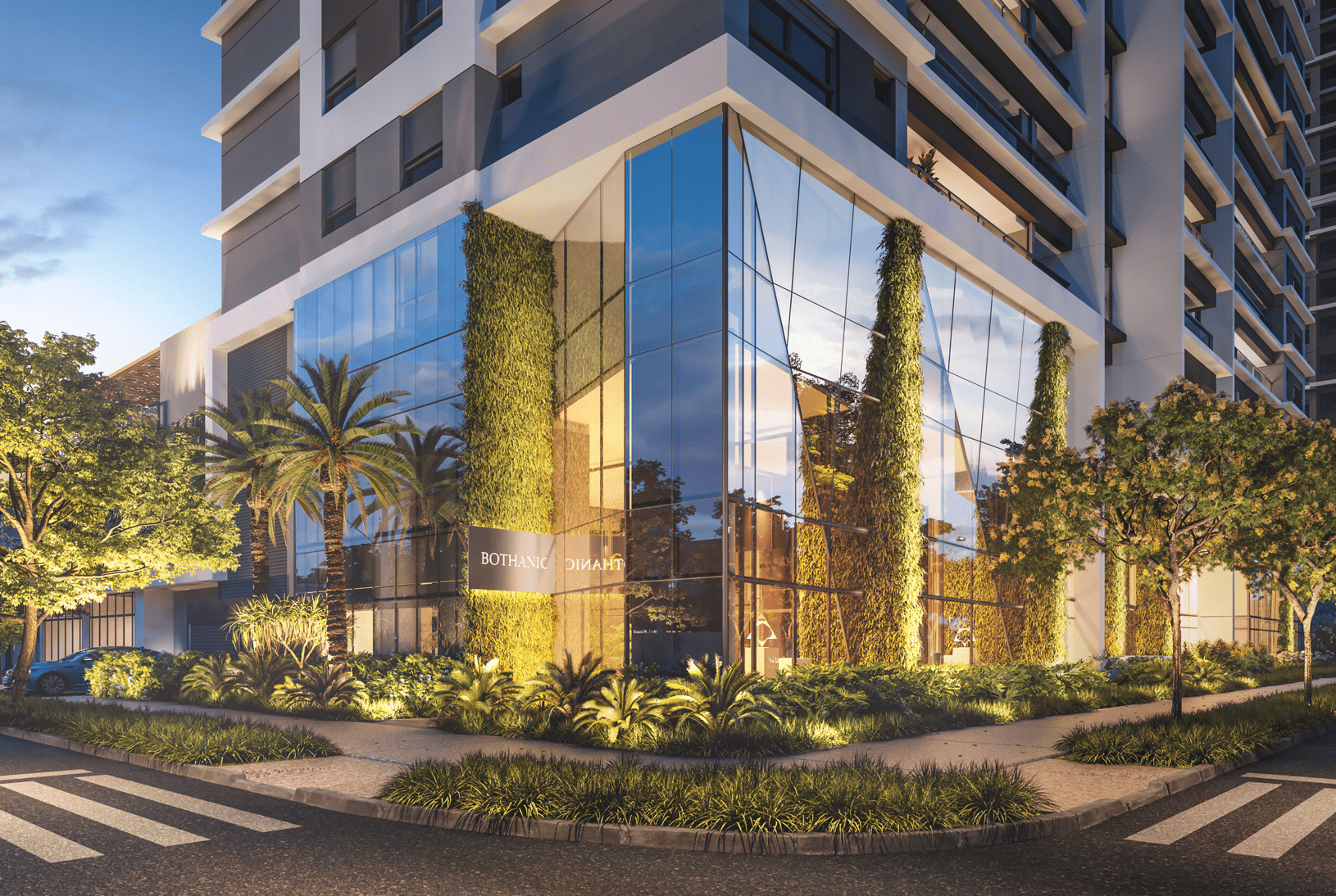 Fachada de vidro iluminada de um edifício moderno com jardins verticais, palmeiras e paisagismo, vista da rua com faixa de pedestres ao anoitecer.